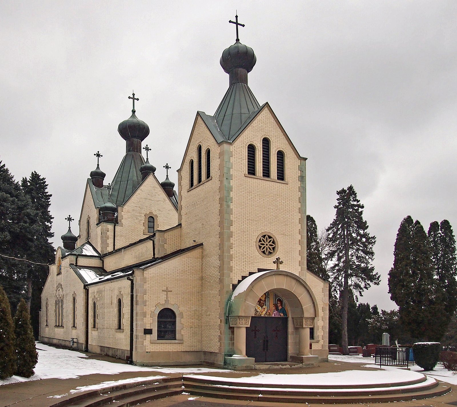 The church of the Monastery of St. Sava in Libertyville, Illinois, a cream brick church with a square bell tower, green copper onion domes and Orthodox crosses, with an icon over the arched entrance and snow on the ground