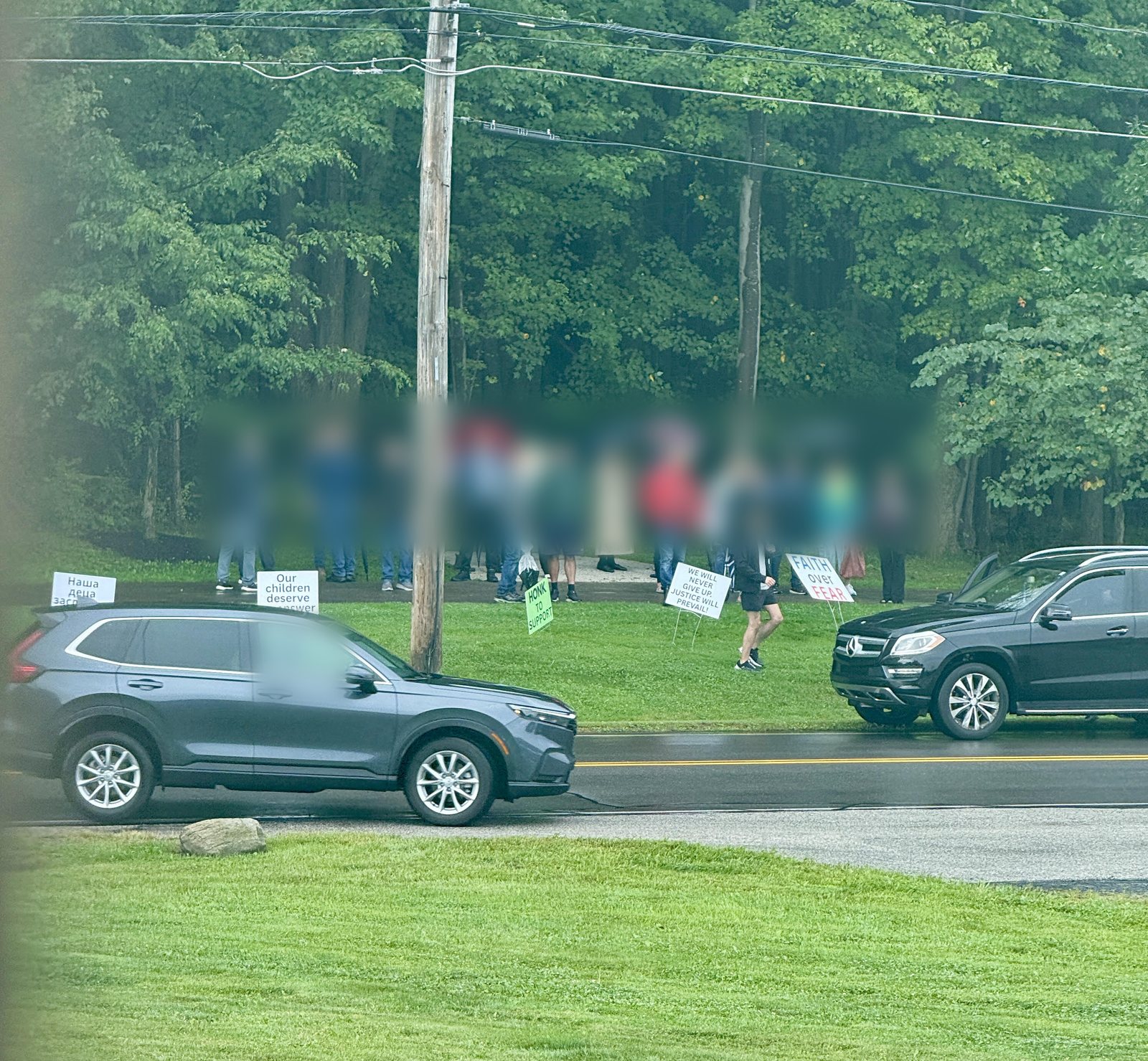 Protesters standing on the grass beside Ridgewood Drive, with signs reading Our children deserve an answer, a matching sign in Serbian, We will never give up, Justice will prevail, Faith over fear, and Honk to support; faces obscured